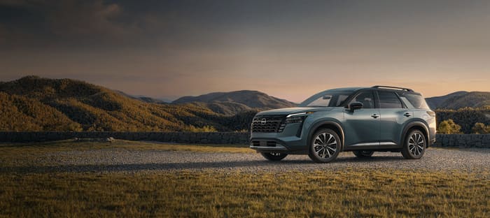 Nissan Pathfinder 2026 en verde azulado, vista de tres cuartos frontal, estacionada sobre gravilla frente a un muro de piedra y montañas al atardecer