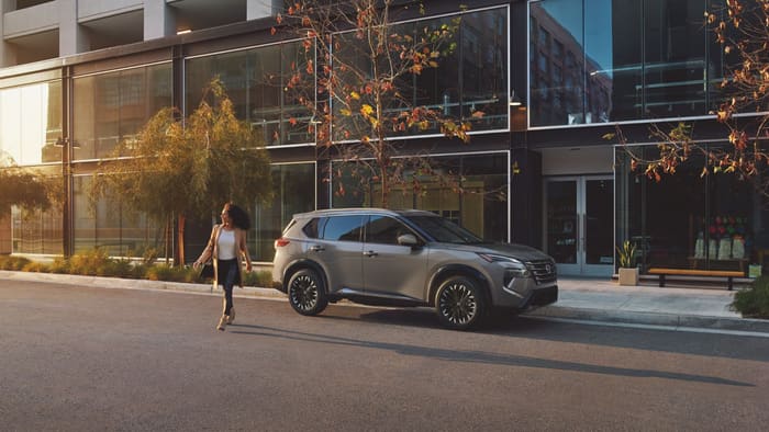 Nissan Rogue 2026 gris en vista tres cuartos delantera, estacionada frente a un edificio de cristal mientras una mujer cruza la calle